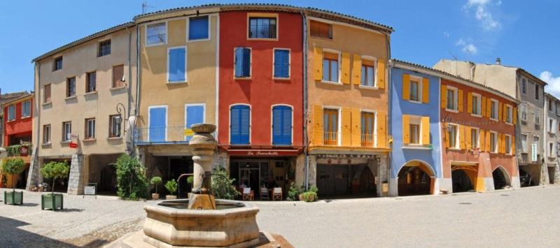 Panoramic photo of the Place des Arcades in Buis-les-Baronnies. In the foreground, the fountain and the cobbled street. In the background, the colorful, adjoining facades: beige, sand, orange-red, light yellow, sky blue, pink, and blue and yellow shutters.